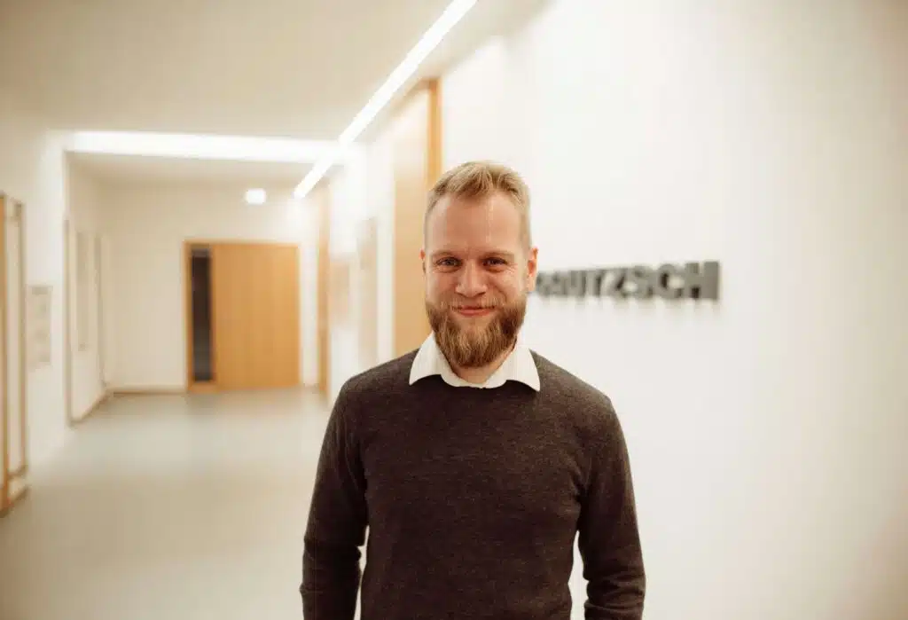 Lächelnder Mann mit Bart, weißem Hemd und grauem Pullover steht in einem hellen Flur mit H. Gautzsch Schriftzug im Hintergrund