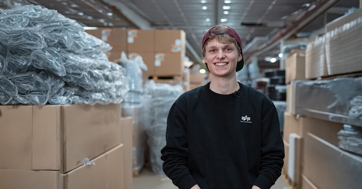 Junger Mann mit Baseballkappe lächelnd, in Lagerhalle, umgeben von verpackten Waren und Kartons