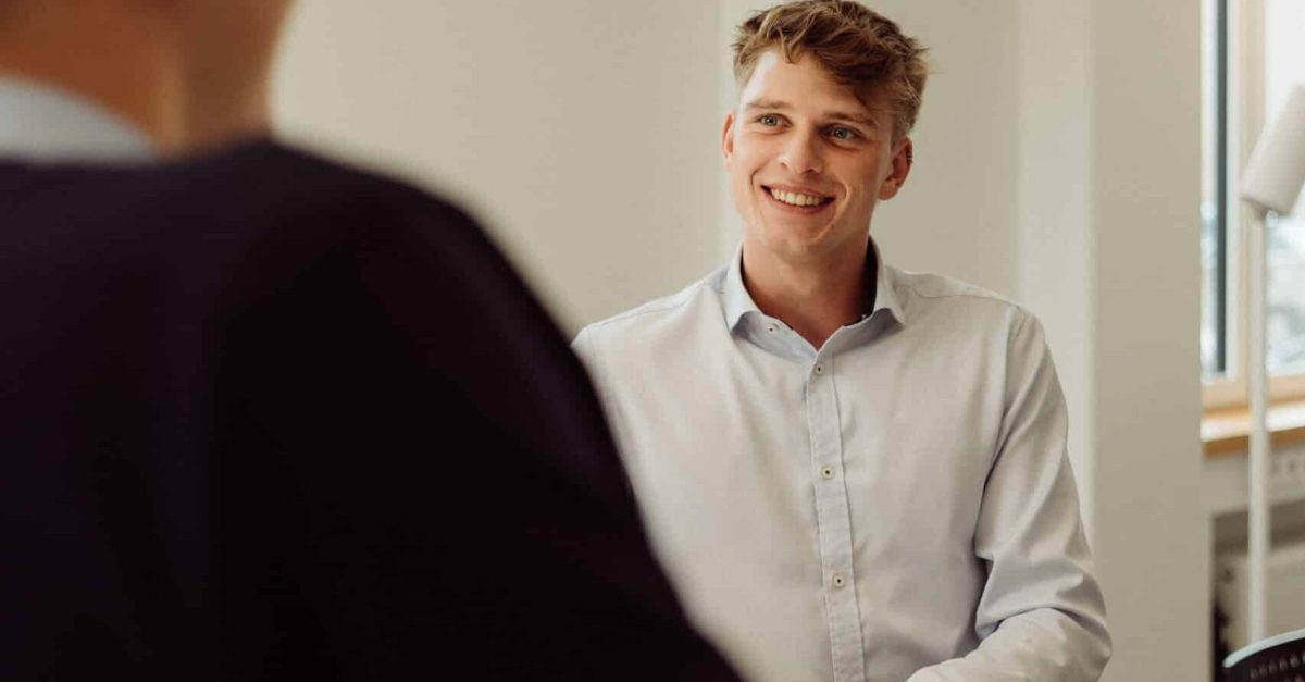 Junger Mann in weißem Hemd lächelt und steht an einem Tisch in einem hellen Büro, während eine andere Person ihm gegenübersteht