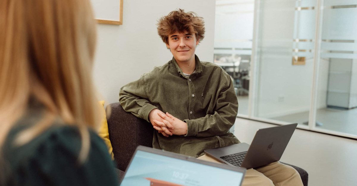 Junger Mann mit lockigem Haar sitzt auf einem Sofa mit Laptop auf dem Schoß, Frau im Vordergrund unscharf