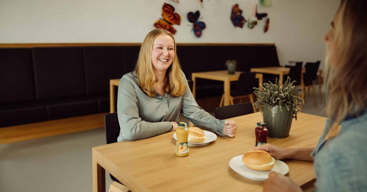 Lächelnde Frau mit langen blonden Haaren sitzt an einem Holztisch in einer Kantine mit bunten Schmetterlingsdekorationen an der Wand