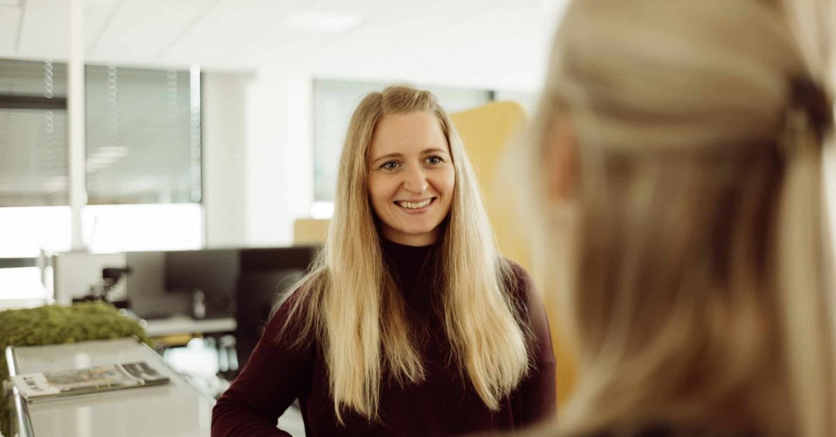 Lächelnde Frau mit langen blonden Haaren steht an einem Empfangstresen und spricht mit einer anderen Frau im Büro