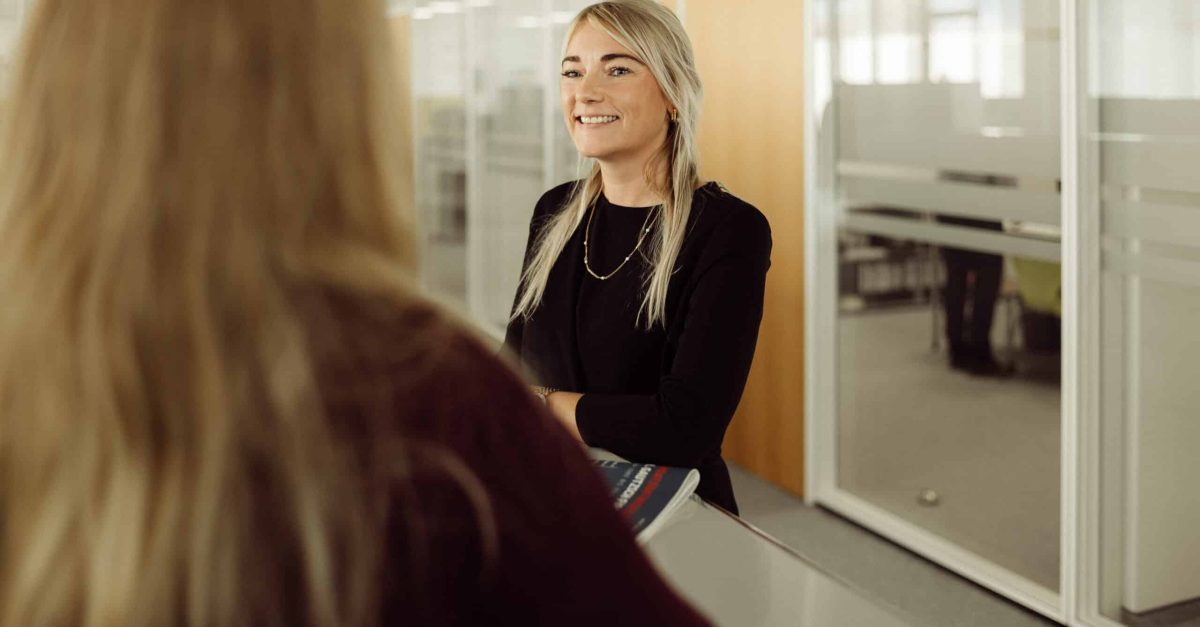 Lächelnde Frau mit blonden Haaren und schwarzem Oberteil steht an einem Empfangstresen in einem Büro