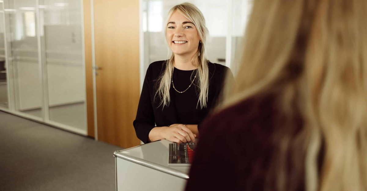 Lächelnde Frau mit blonden Haaren und schwarzem Oberteil steht an einem Empfangstresen in einem Büro
