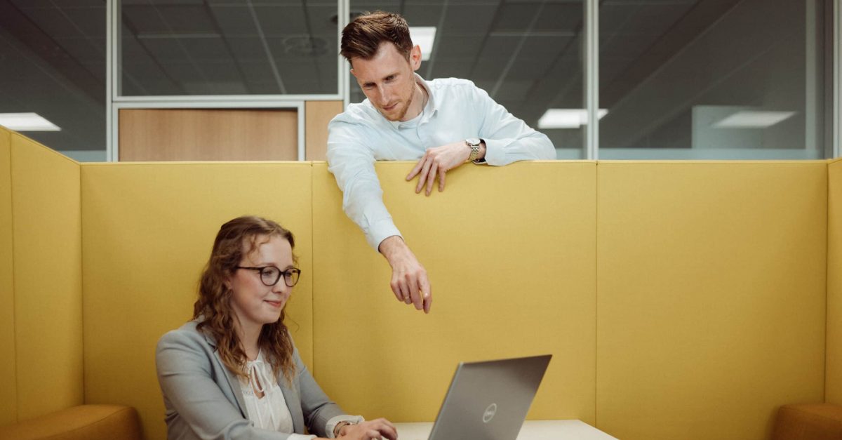 Frau sitzt an einem Laptop in einem Büro, Mann steht hinter einer gelben Trennwand und zeigt auf den Bildschirm