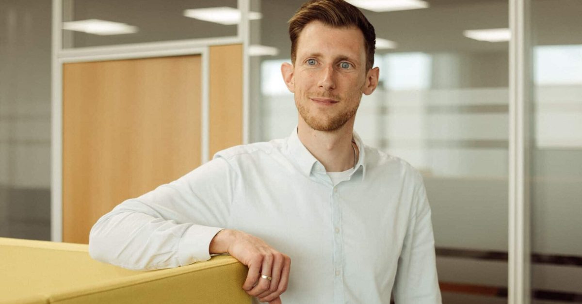 Junger Mann mit weißem Hemd und Jeans lehnt lächelnd an gelber Bürowand in modernem Büro