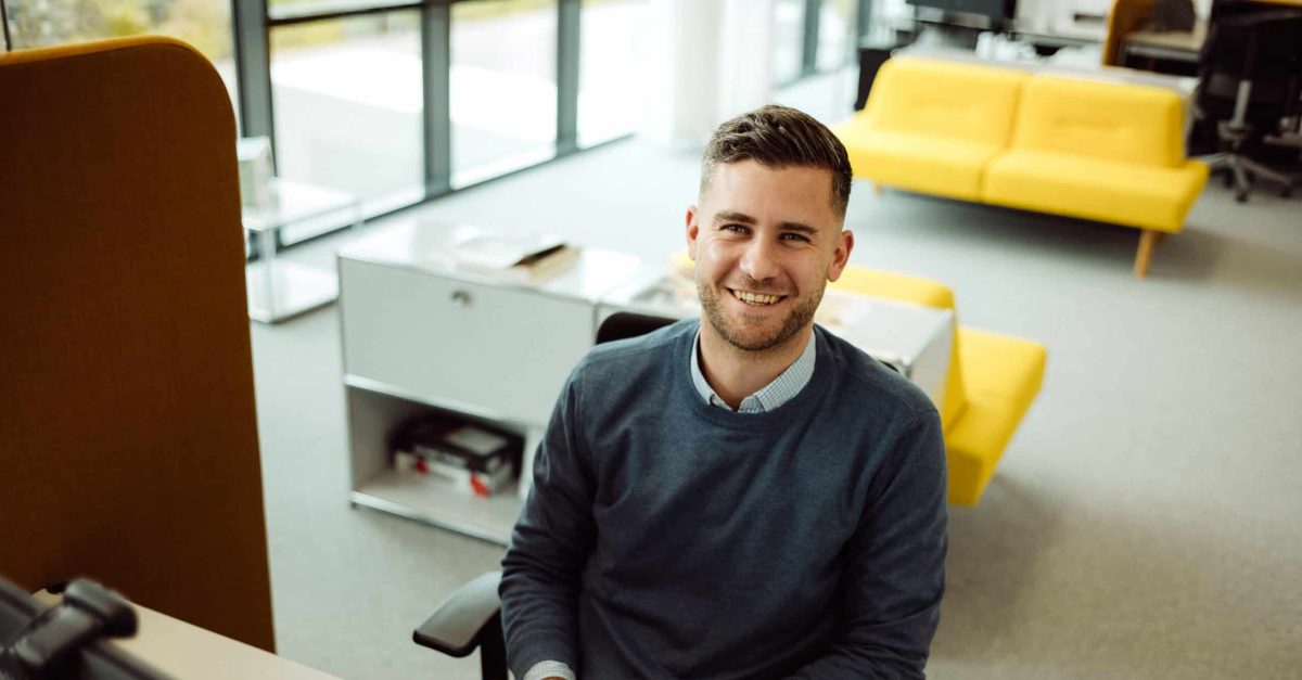 Lächelnder Mann mit kurzen braunen Haaren sitzt an einem Schreibtisch im Büro vor mehreren Monitoren