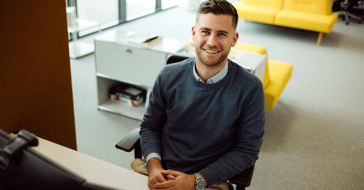 Lächelnder Mann mit kurzen braunen Haaren sitzt an einem Schreibtisch im Büro vor mehreren Monitoren
