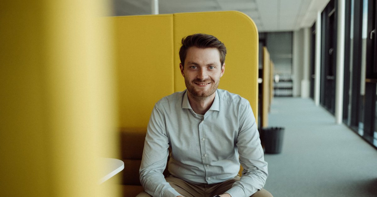 Junger Mann mit Hemd und Uhr sitzt lächelnd in einem modernen Büro mit gelben Trennwänden