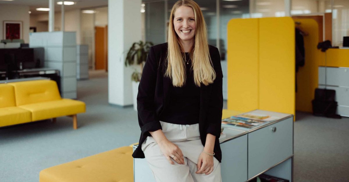 Lächelnde Frau mit langen blonden Haaren, schwarzem Oberteil und weißer Hose, sitzt in modernem Büro mit gelben Möbeln