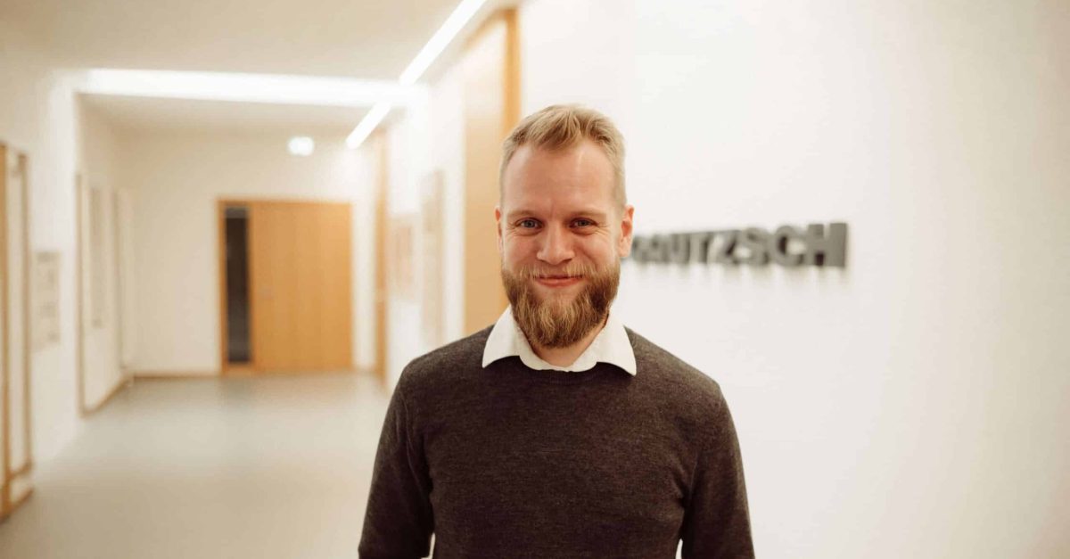Lächelnder Mann mit Bart, weißem Hemd und grauem Pullover steht in einem hellen Flur mit H. Gautzsch Schriftzug im Hintergrund