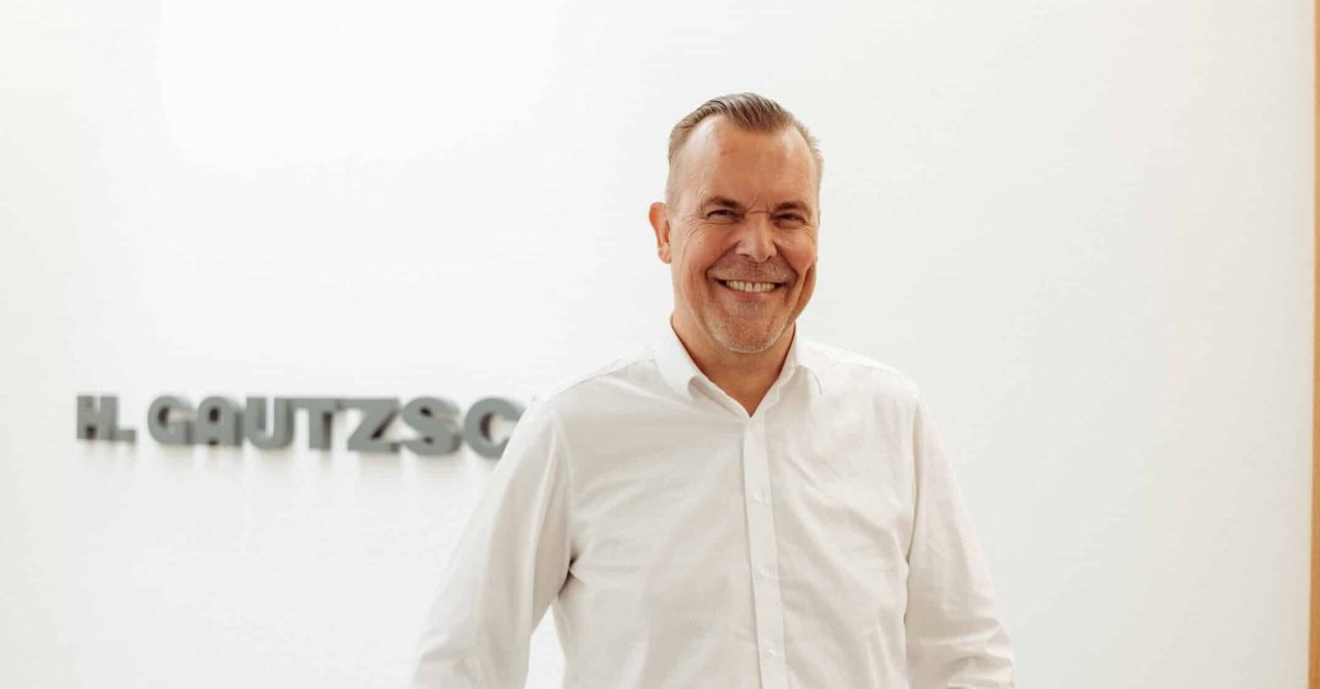 Lächelnder Mann mit weißem Hemd steht angelehnt in einem hellen Flur mit H. Gautzsch Schriftzug im Hintergrund