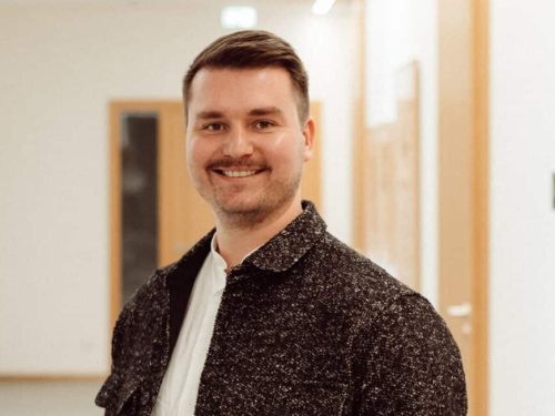 Lächelnder Mann mit weißem Hemd und dunkler Jacke steht in einem hellen Flur mit H. Gautzsch Schriftzug im Hintergrund