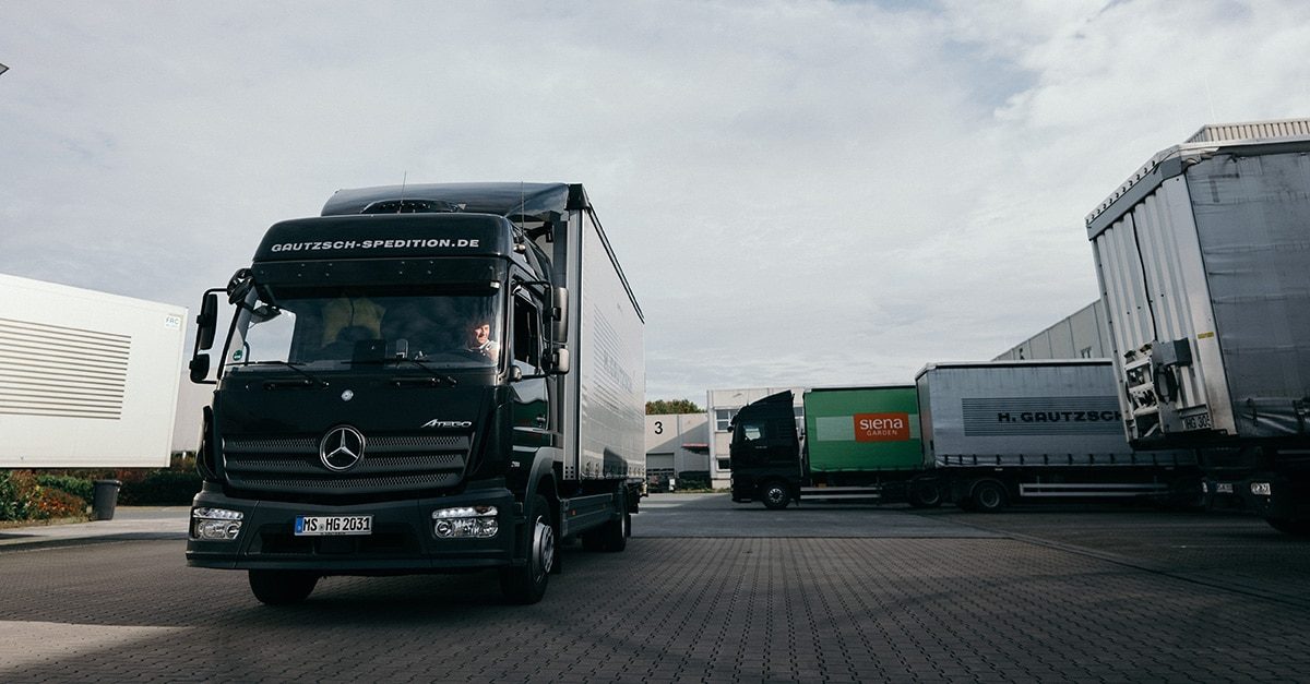 Schwarzer Lkw fährt auf einem Industriehof mit weiteren geparkten Lkw und Lagerhallen im Hintergrund