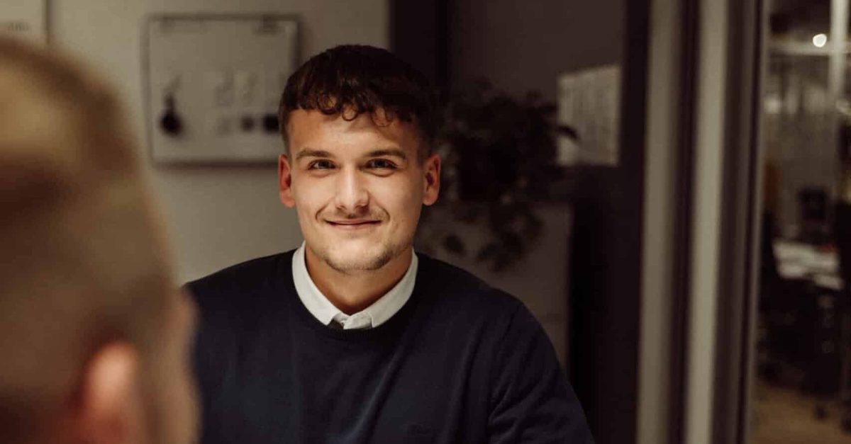 Junger Mann mit dunklem Pullover und weißem Hemd sitzt lächelnd an einem Tisch in einem modernen Büro