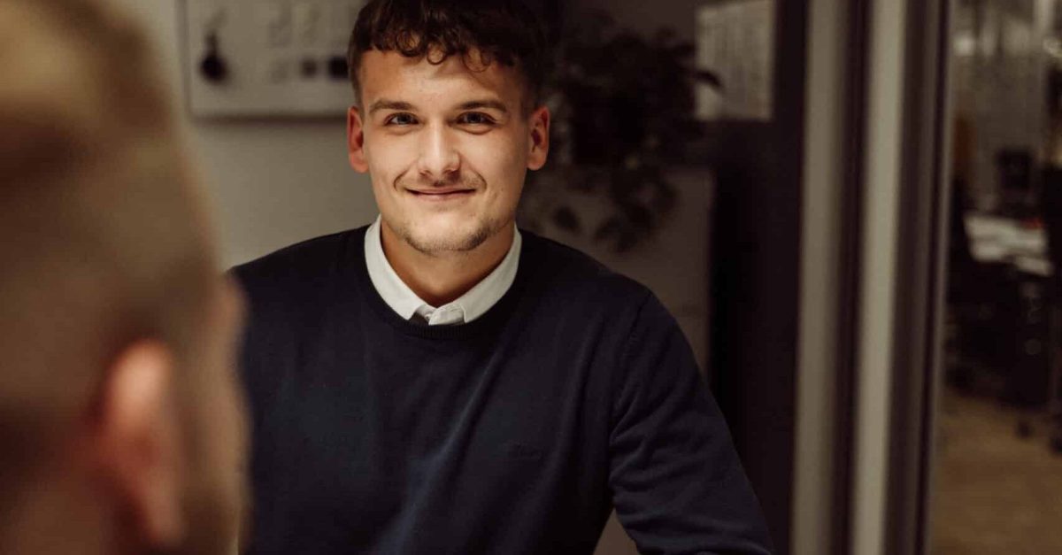 Junger Mann mit dunklem Pullover und weißem Hemd sitzt lächelnd an einem Tisch in einem modernen Büro