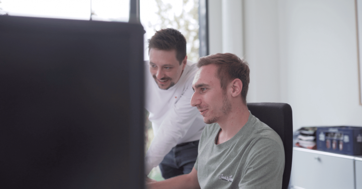 Zwei Männer arbeiten gemeinsam an einem Computer in einem hellen Büro, einer sitzt und der andere steht und schaut lächelnd auf den Bildschirm.
