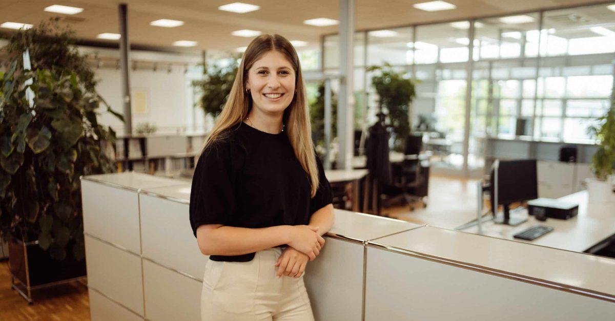Junge Frau mit langen Haaren lächelt stehend in einem modernen Büro mit Schreibtischen und Pflanzen